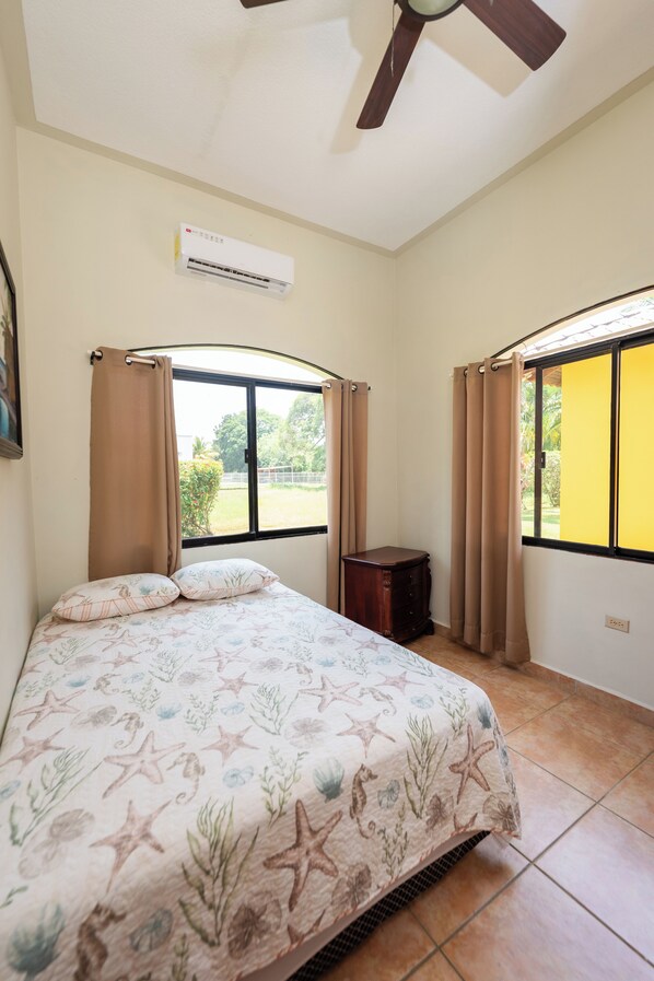 Un dormitorio con cama, aire acondicionado, mesita de noche y ventana con cortinas.