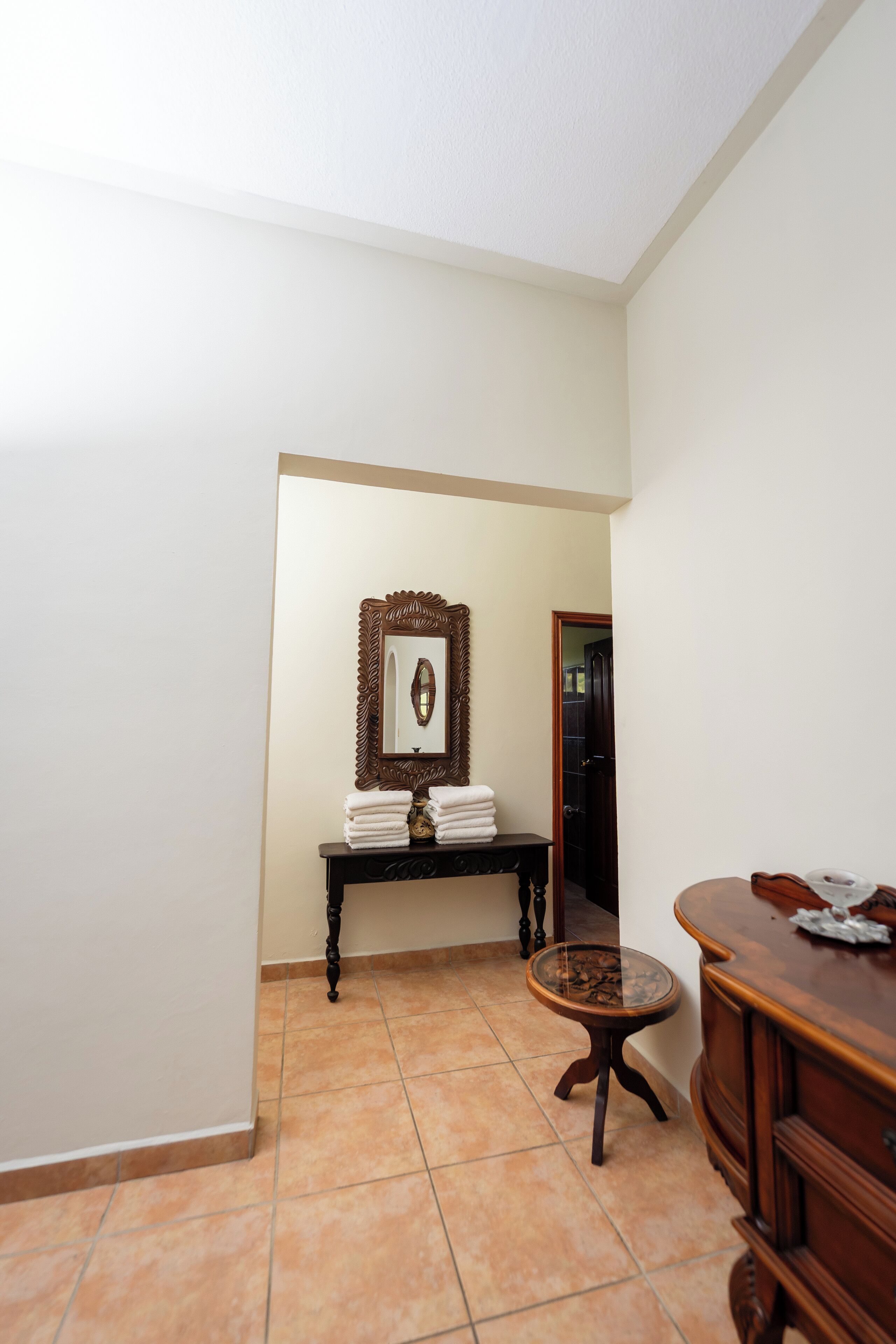 A hotel room with a tiled floor, a wooden dresser, a small round table, and a mirror with an ornate frame.