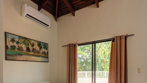 Een slaapkamer met een bed, een airconditioning, een schilderij en uitzicht door een raam met gordijnen.
