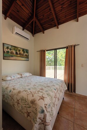 Une chambre à coucher avec un lit, un appareil de climatisation, un tableau et une vue par la fenêtre, qui est ornée de rideaux.