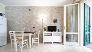 Um espaço de estar compacto com parede de pedra, mesa de jantar com cadeiras, televisão em um armário branco e uma porta de vidro deslizante à direita.