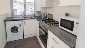 A compact kitchen with a washing machine, a gas stove, a microwave, and a window with blinds. - Pinecone Lodge - No 9 (Morpeth)