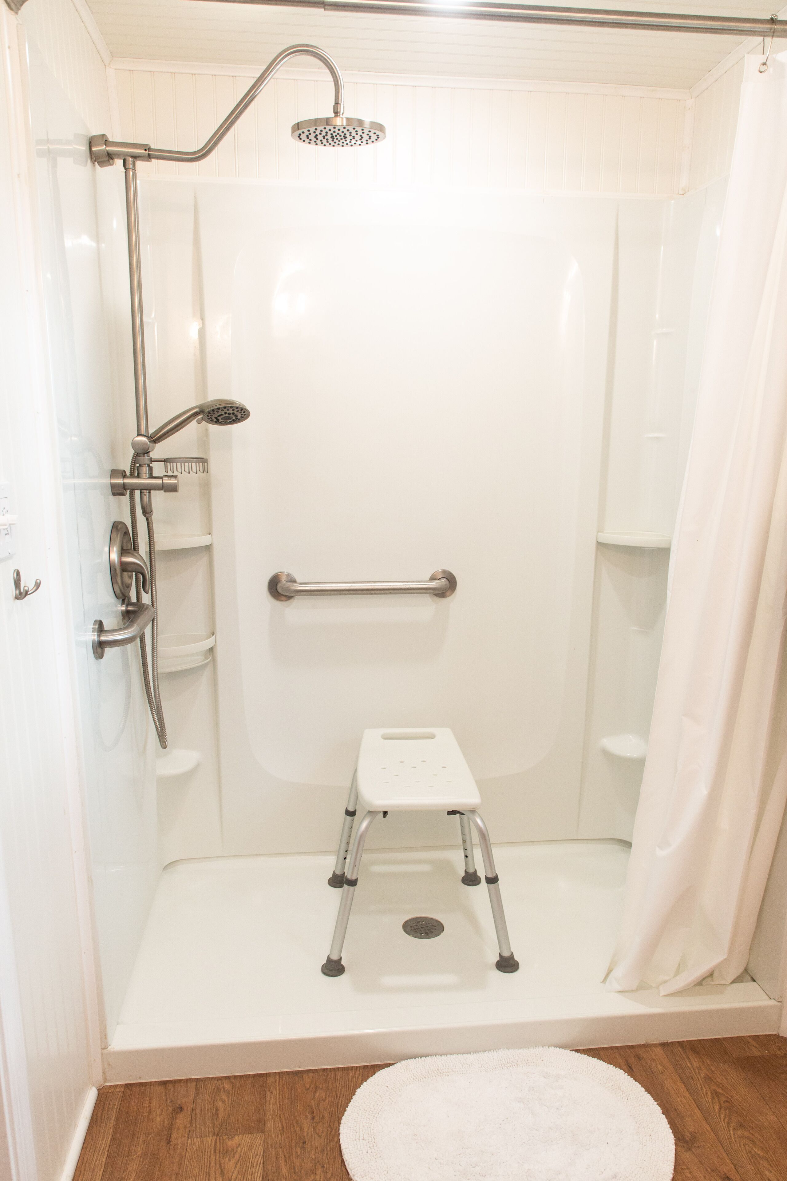 Doccia accessibile con porte scorrevoli, barra di sostegno, soffione e un piccolo sgabello bianco.