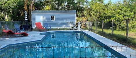 A swimming pool with a wire fence, lounge chairs, and a small building in the background.