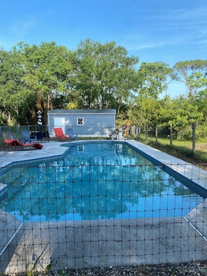 A swimming pool with a wire fence, lounge chairs, and a small building in the background.
