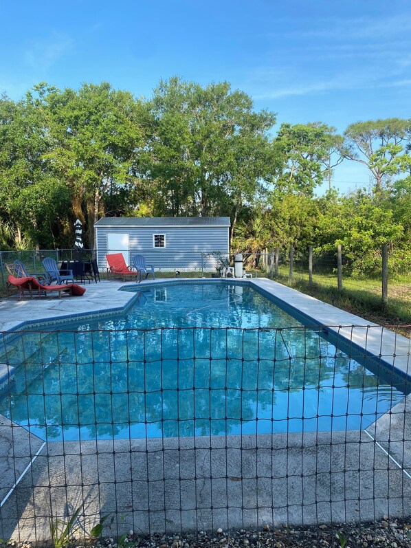 A swimming pool with a wire fence, lounge chairs, and a small building in the background.