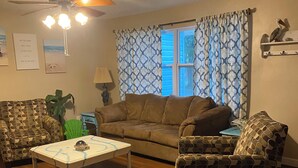 Un salón con un sofá estampado, una mesa de centro, un ventilador de techo y una ventana con cortinas estampadas.