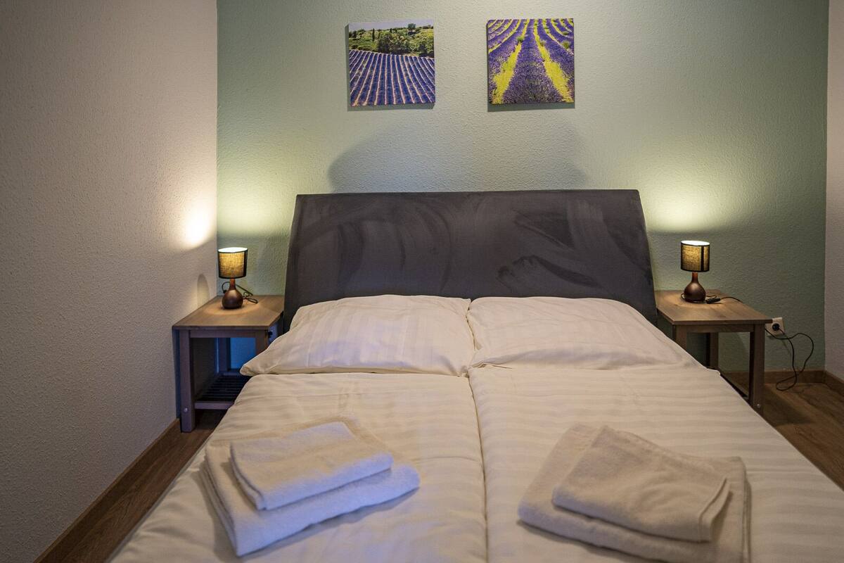 A neatly made bed with folded towels, two bedside lamps, and two paintings on the wall.