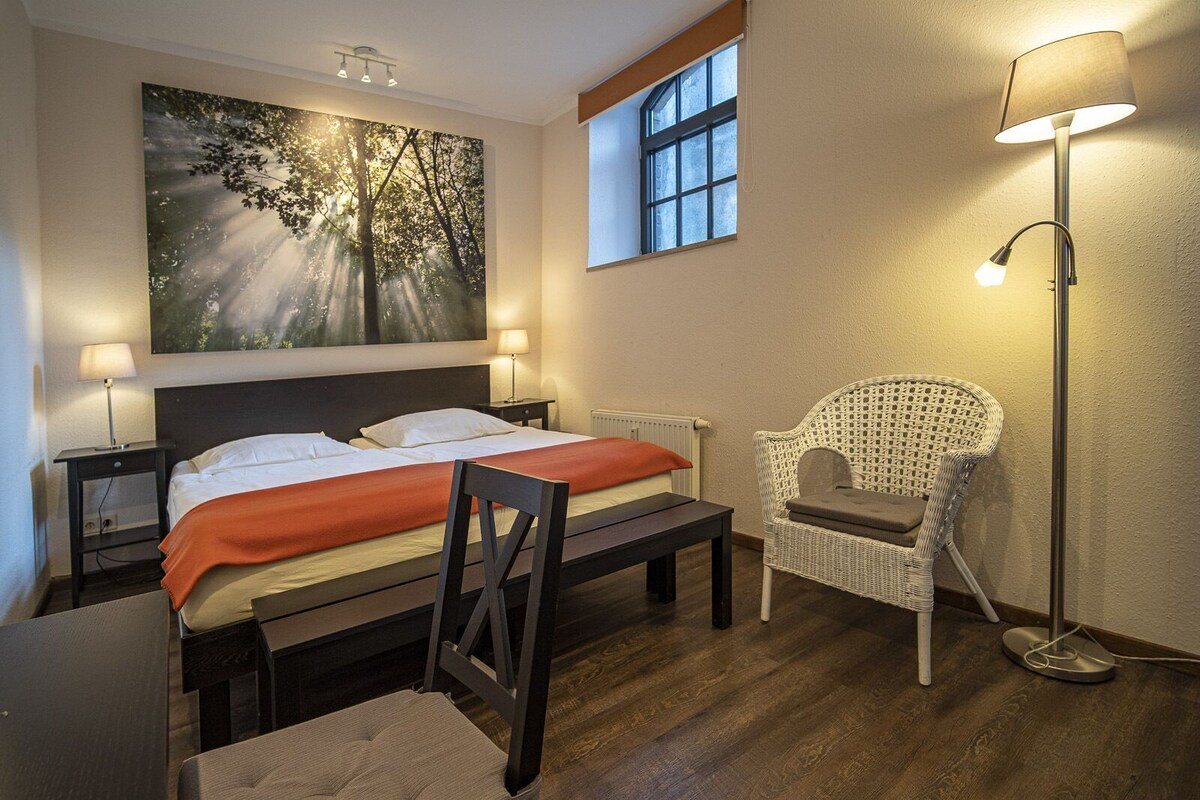Una habitación de hotel con una cama, dos mesitas de noche con lámparas, una silla, un ventanal grande y una obra de arte en la pared.