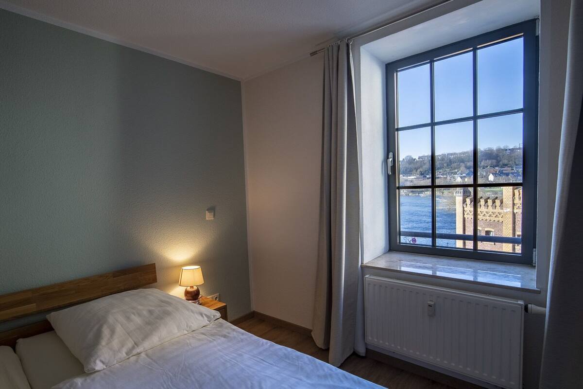 A bedroom with a large window, a bed, a nightstand, and a lamp.
