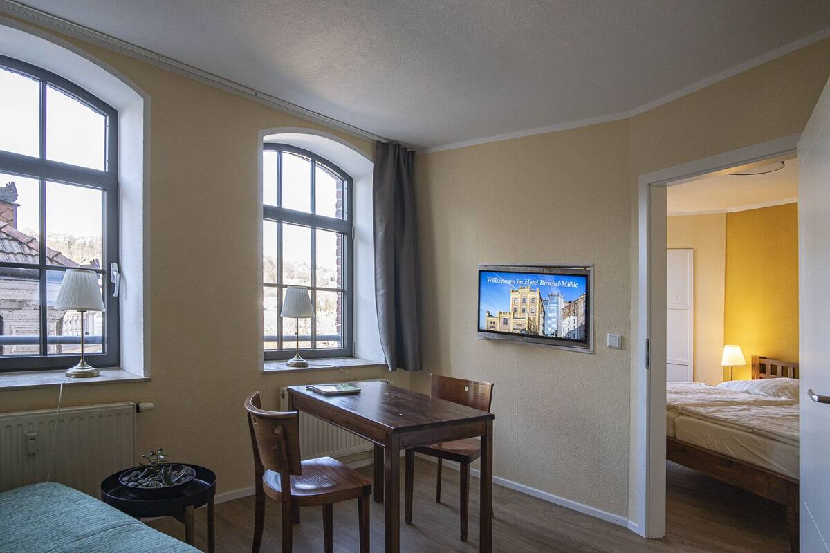 A hotel room with a bed, a desk with a chair, a small table, a radiator, and a TV on the wall.