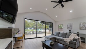 A modern living room with a large window, a ceiling fan, a flat-screen TV, a grey sofa, and a dining area with yellow chairs. - One of a kind home in the city (Arlington)