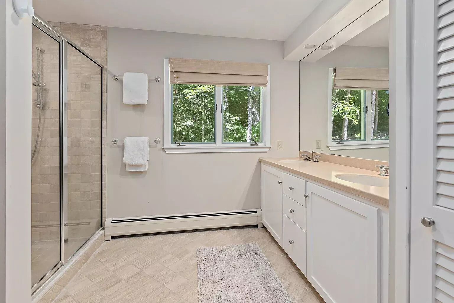 Un baño moderno con cabina de ducha de vidrio, un espejo grande, un tocador color beige con lavamanos y una ventana que permite la entrada de luz natural.