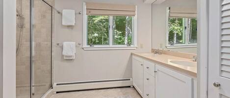 Un baño moderno con cabina de ducha de vidrio, un espejo grande, un tocador color beige con lavamanos y una ventana que permite la entrada de luz natural.