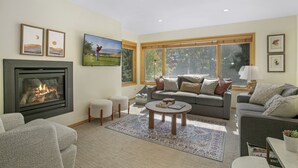 A living room with a fireplace, a grey sofa, a coffee table, and a large window with a view of trees. - Modern Vail Retreat, Free Shuttle, Fireplace & Mountain Views | SF201 (Vail)