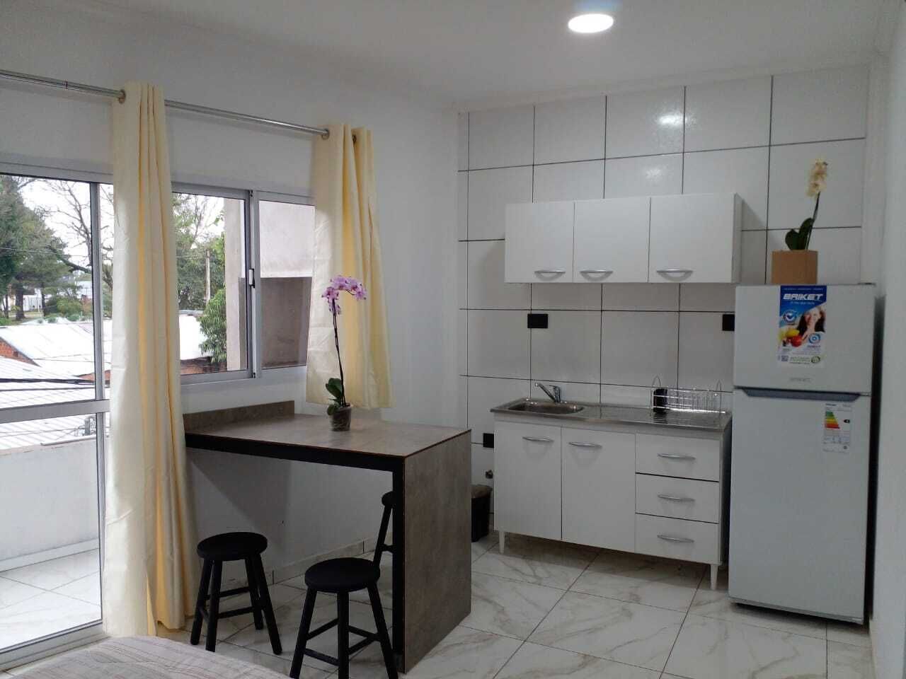 Una cocina moderna con armarios blancos, un lavamanos empotrado y un refrigerador. Se aprecia una pequeña isla con taburetes y una planta en maceta.