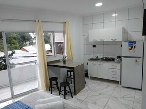 A modern kitchen with white cabinets, a built-in stove, and a refrigerator. There are two stools and a window with curtains.