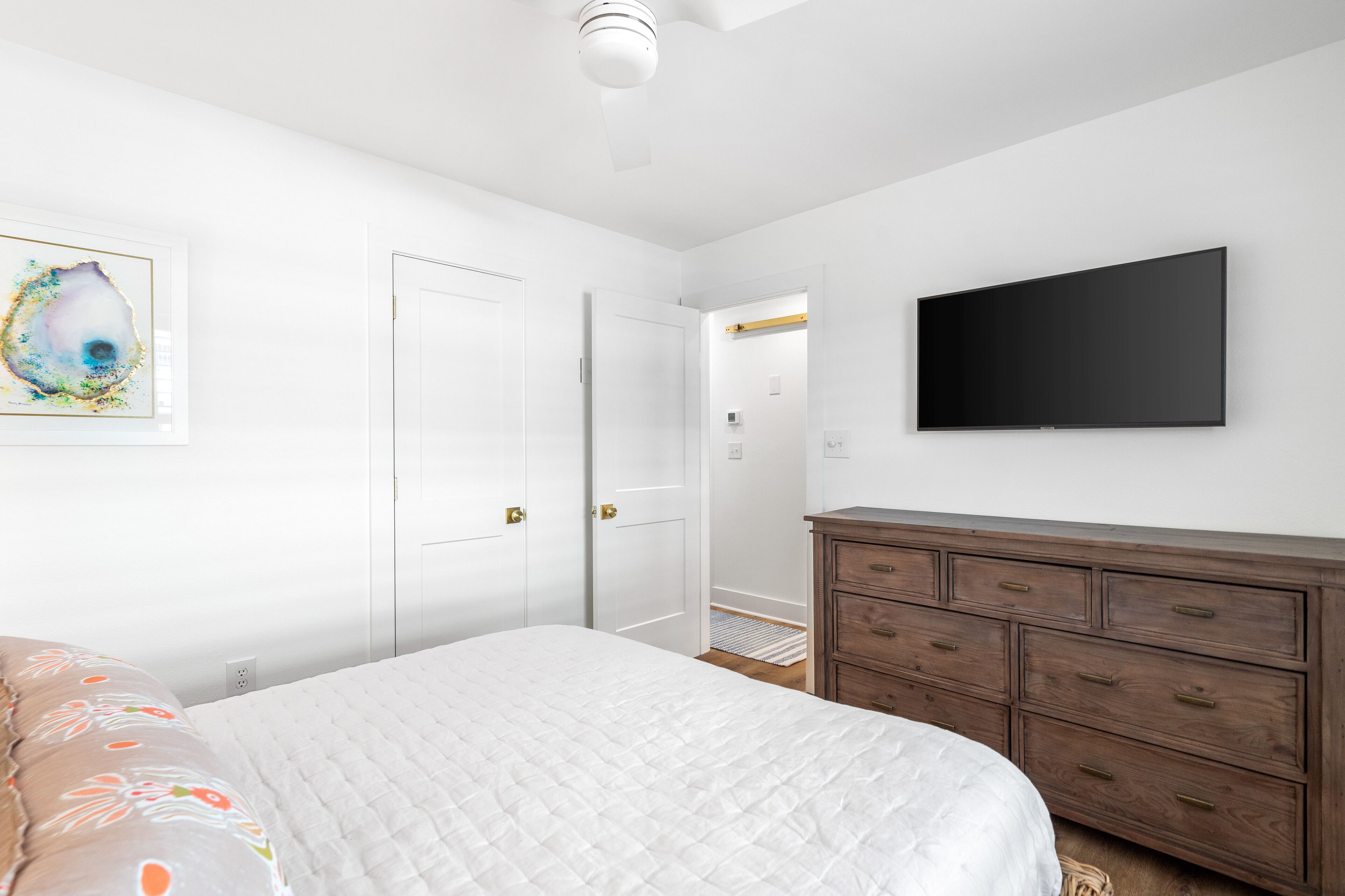 A bedroom with a bed, a dresser, a flat-screen TV, and a door leading to another room.