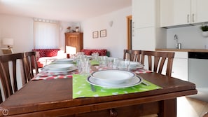 A dining table set with plates, glasses, and cutlery, surrounded by wooden chairs. A kitchen area is visible in the background.