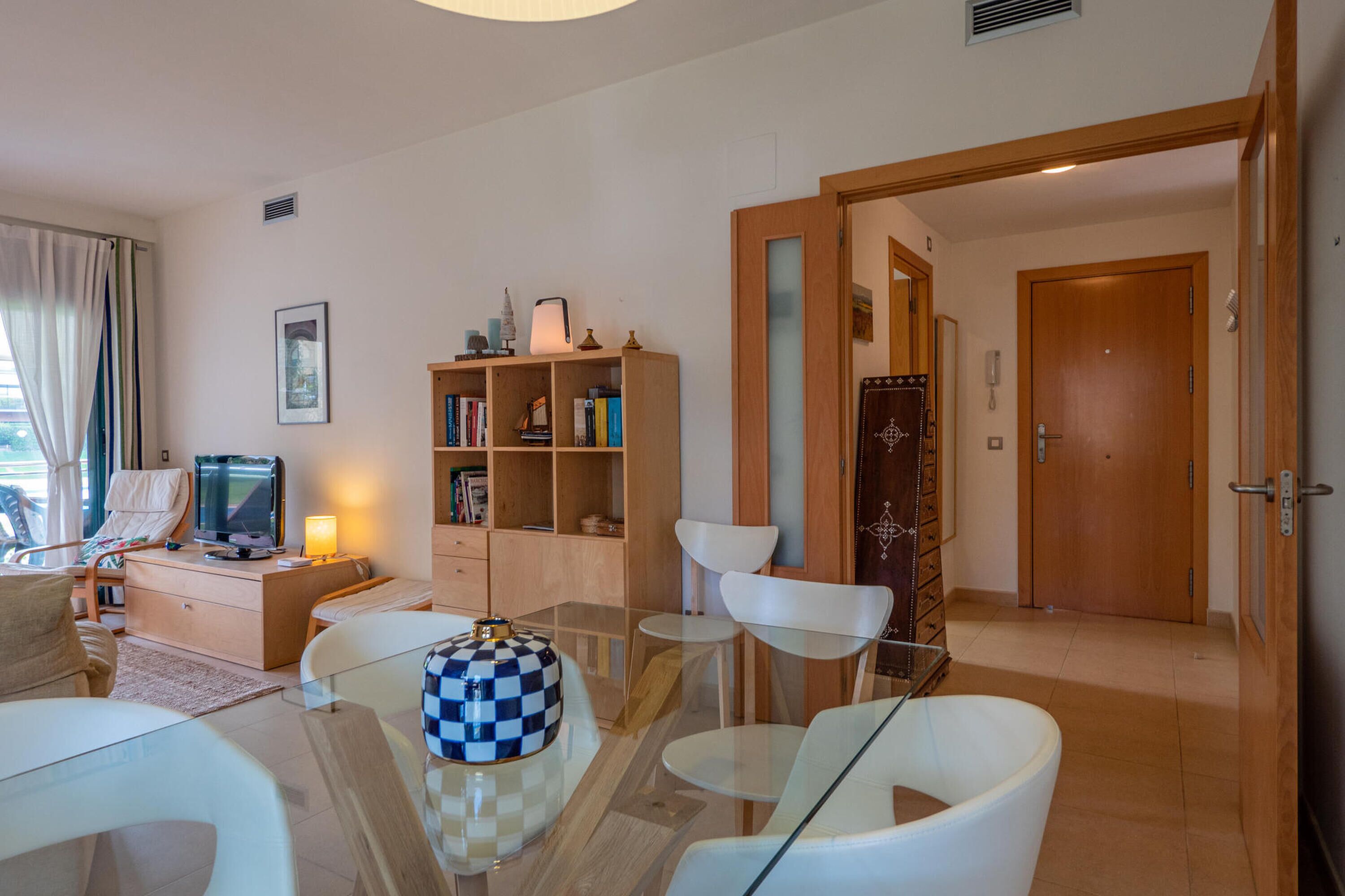 Una sala de estar moderna con una mesa de centro de vidrio, sillas blancas, una estantería y un televisor.