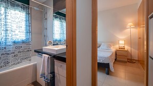 A modern bathroom with a shower, sink, and mirror. There is a bed with a nightstand and lamp in the adjacent bedroom. - Apartment in Pals with community pool (Pals)