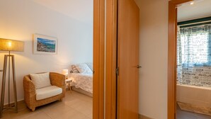 A room with a wicker sofa, a bedside table with a lamp, a bed with white bedding, and a door leading to a bathroom with patterned tiles. - Apartment in Pals with community pool (Pals)