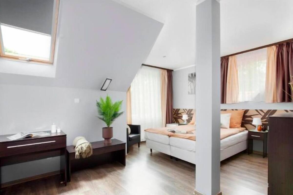 Una habitación moderna y luminosa con escritorio, sofá y cama. Cuenta con una ventana con cortinas, una planta en maceta y un cuadro en la pared.