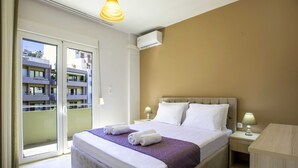Habitación de hotel con cama, mesitas de noche, lámpara, aire acondicionado y un balcón con vistas a otros edificios.