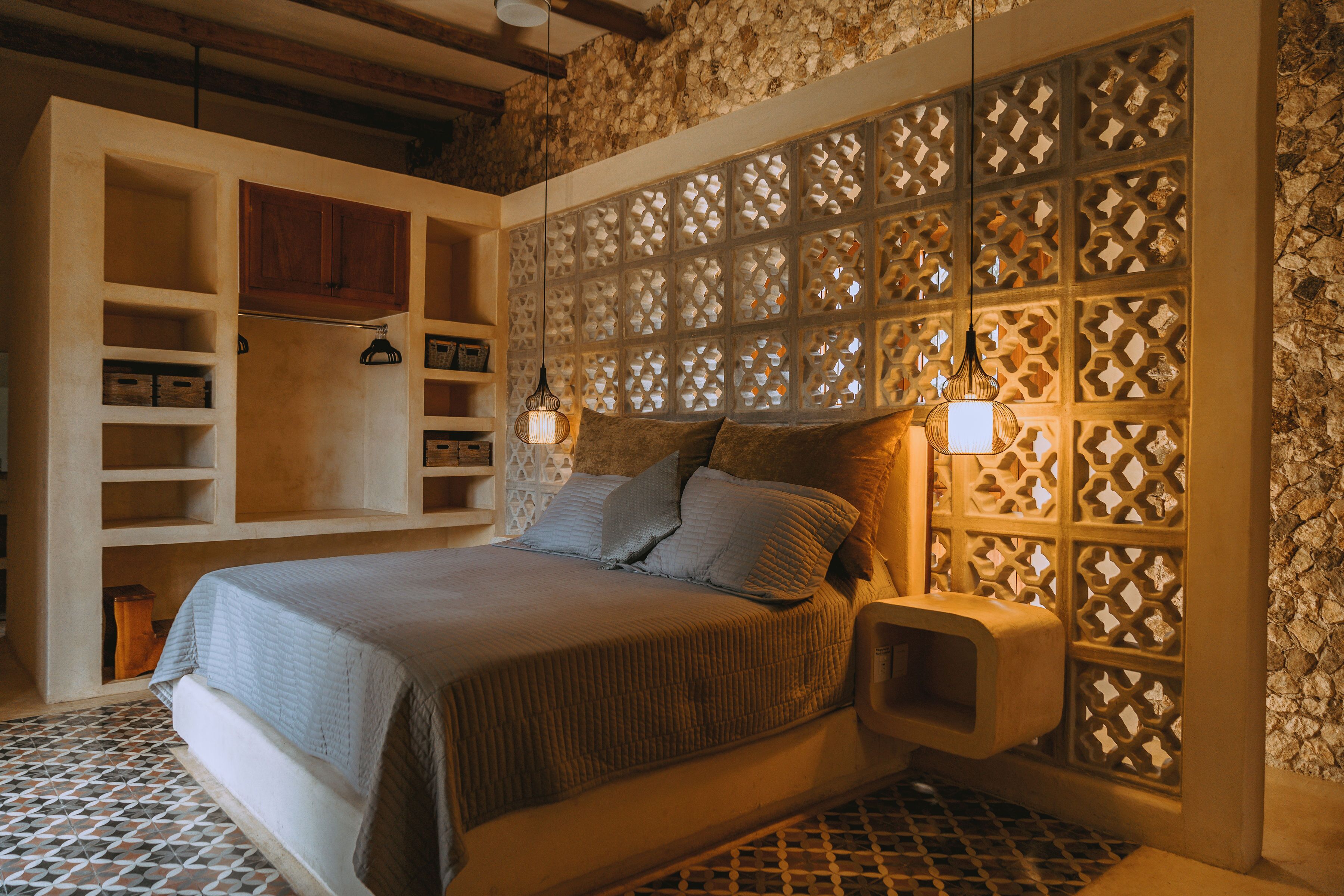 A bedroom with a bed, built-in shelving, a decorative wall panel, and a small bedside table with a lamp.