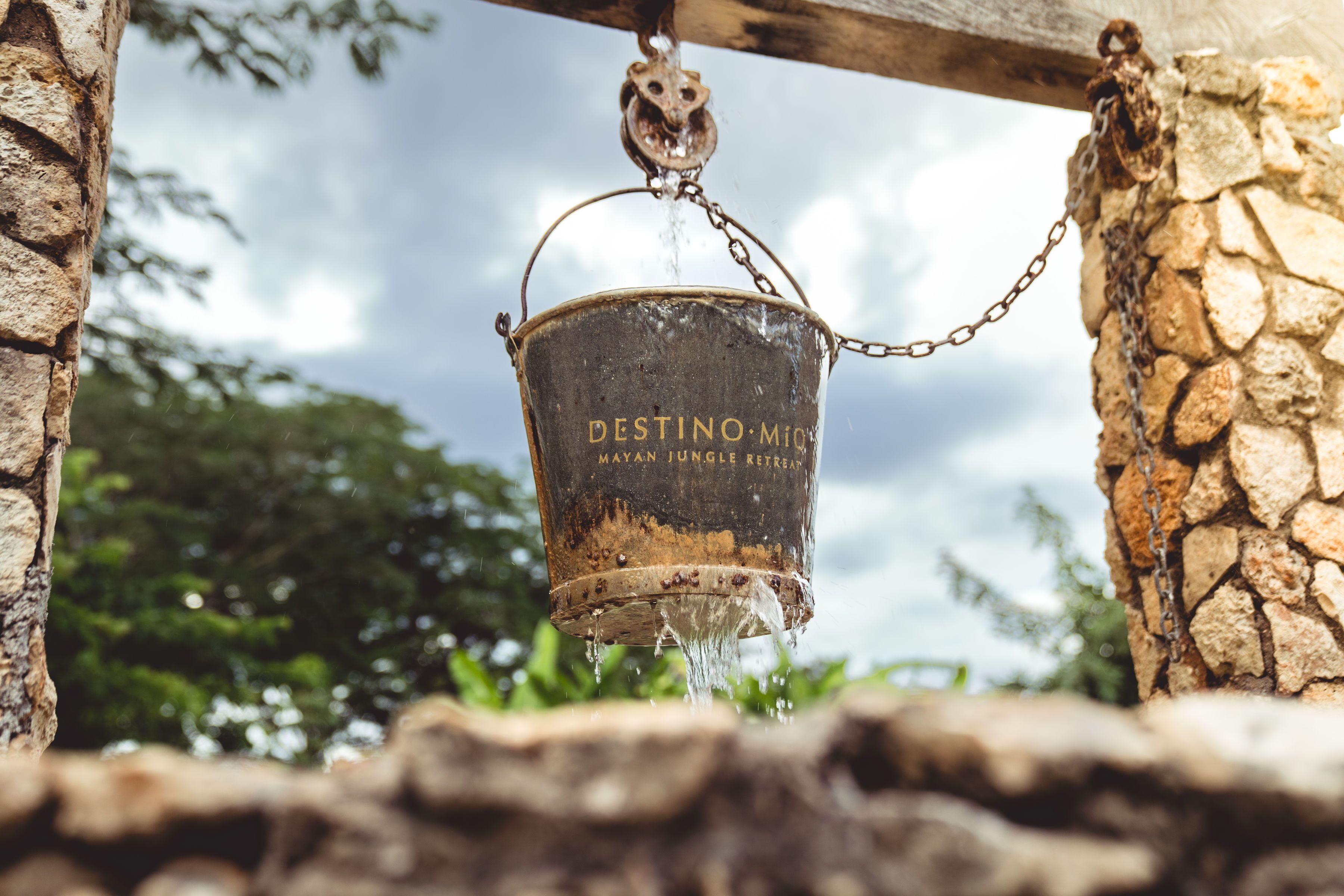 Een robuuste metalen emmer met ketting, voorzien van de tekst “DESTINO MIJ”, hangend aan een houten balk.