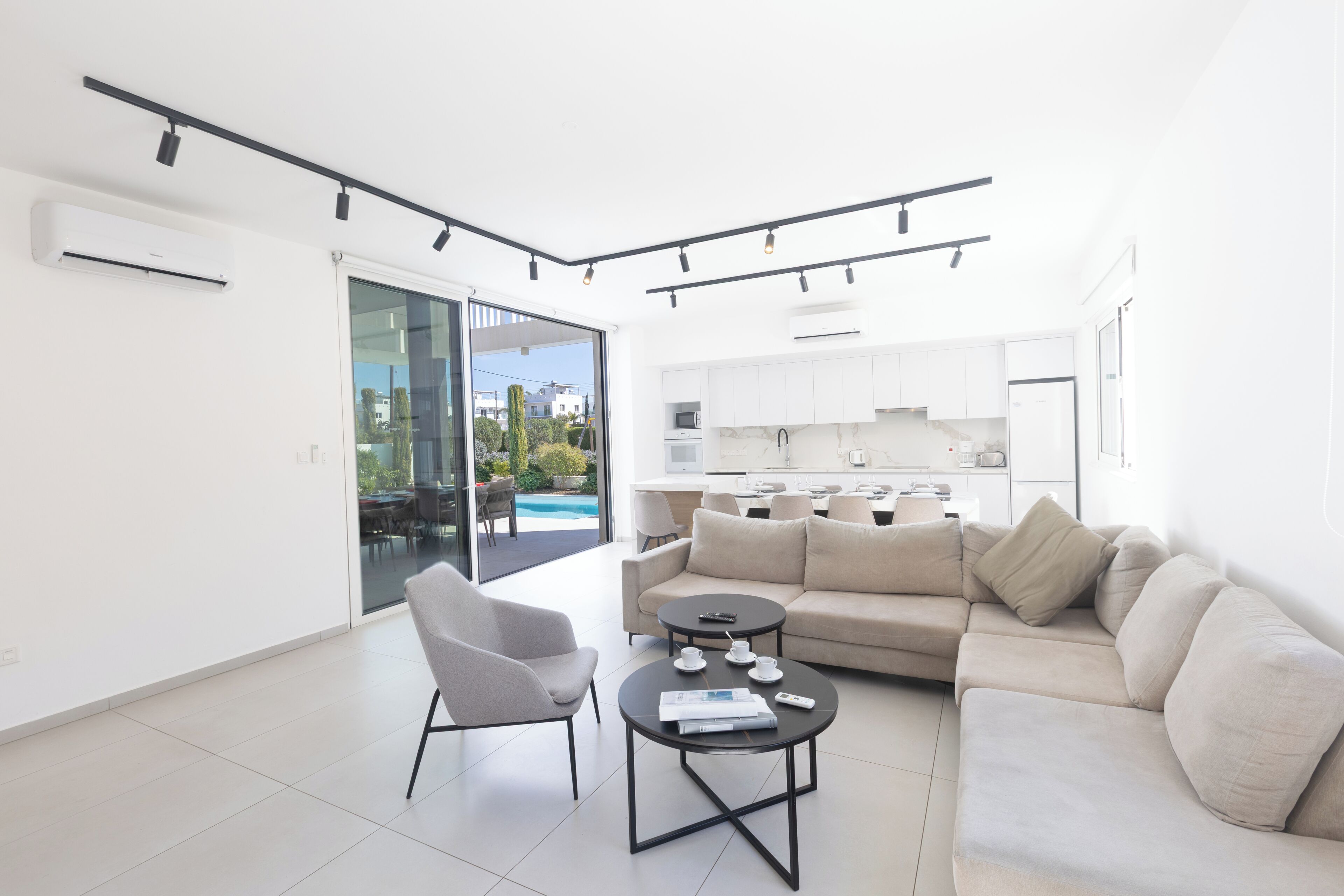 A modern living room with a sofa, armchair, and coffee table, a kitchen in the background, and a sliding glass door leading to an outdoor area.