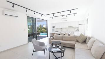 A modern living room with a sofa, armchair, and coffee table, a kitchen in the background, and a sliding glass door leading to an outdoor area.