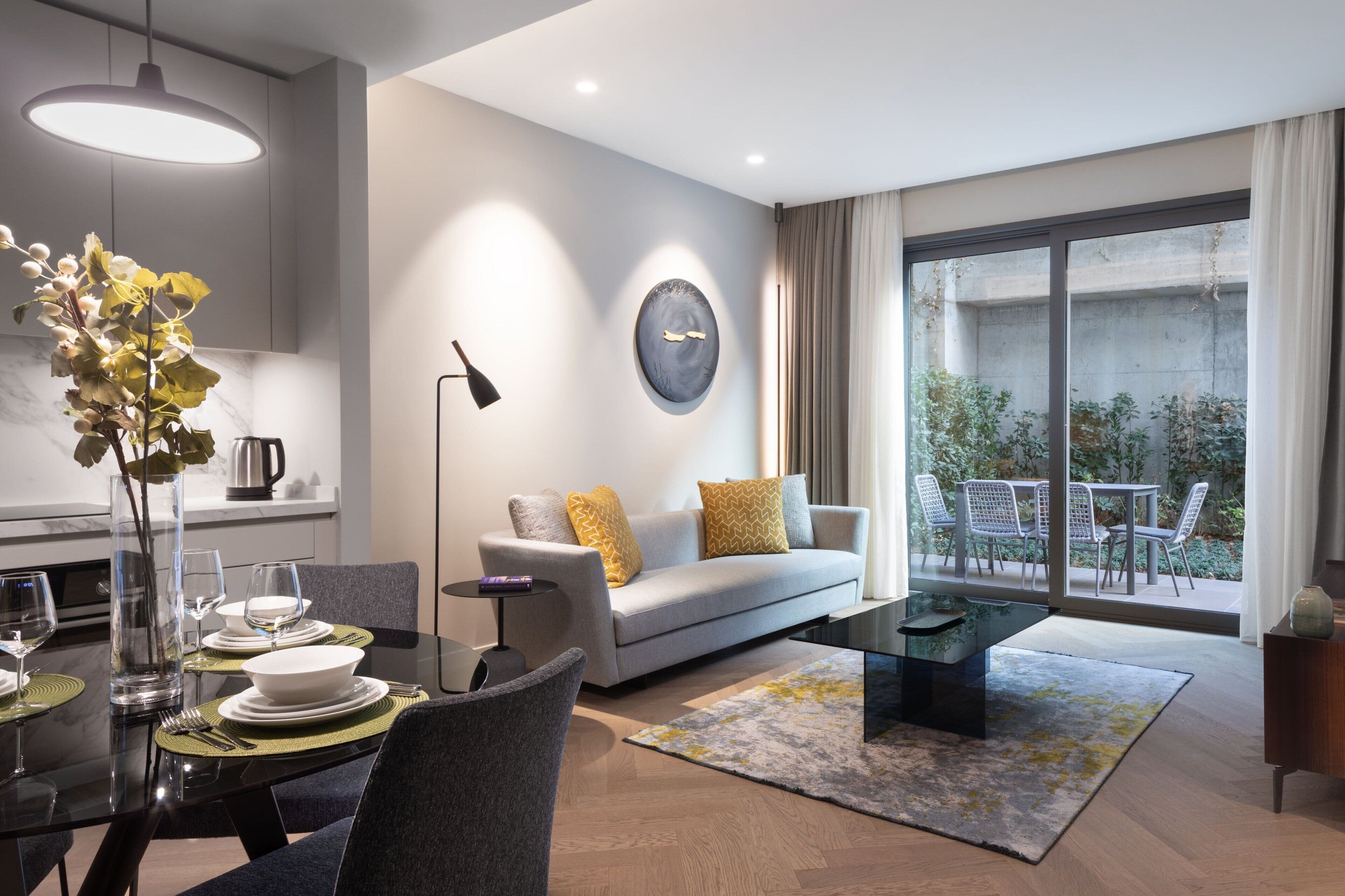 A modern living room with a sofa, coffee table, and dining area. There is a large window with a view of greenery outside.