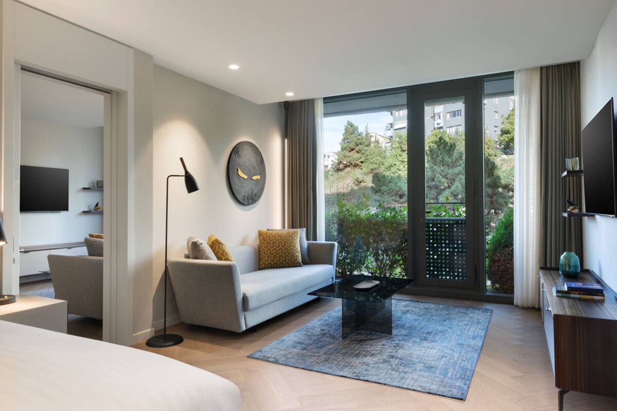 A modern hotel room with a sofa, a coffee table, a TV, and a balcony with a view of greenery and buildings.