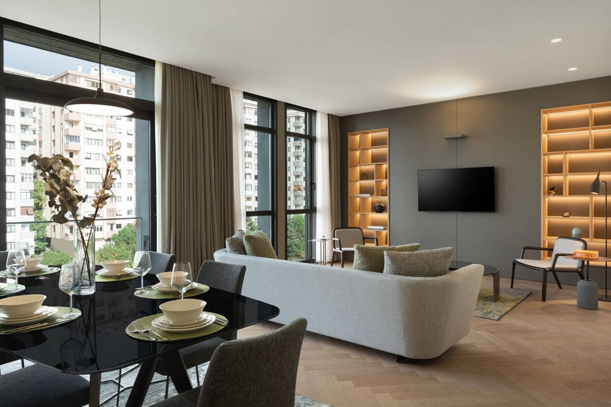 A modern living room with a sofa, dining table, and chairs. There is a TV mounted on the wall and a large window with curtains.