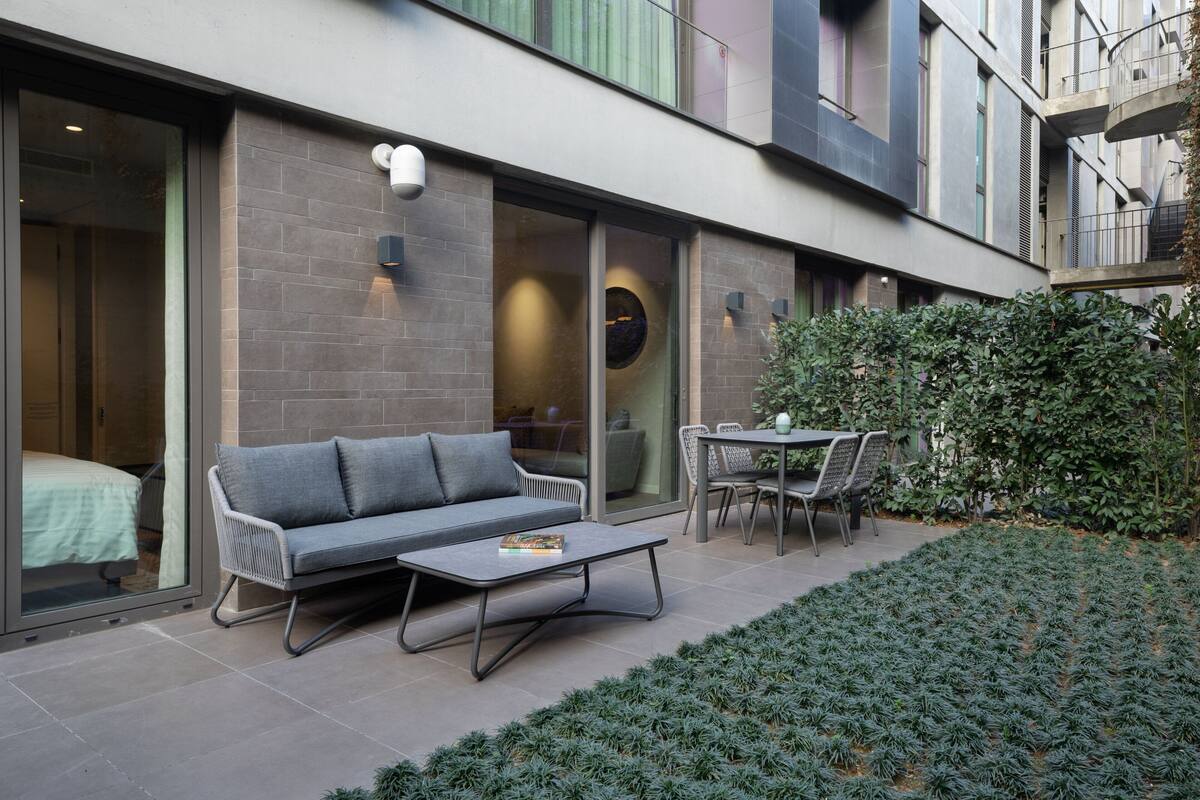 A modern outdoor seating area with a sofa and table, a small garden, and a building with large windows and a balcony.