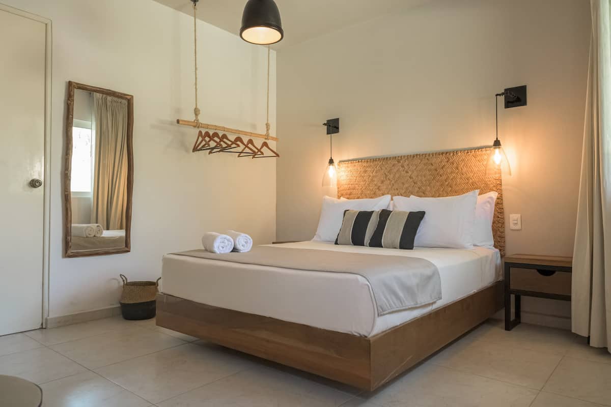 Una cama bien hecha con sábanas blancas y dos toallas enrolladas, un cabecero de madera, una lámpara colgante y una mesita de noche con lámpara.