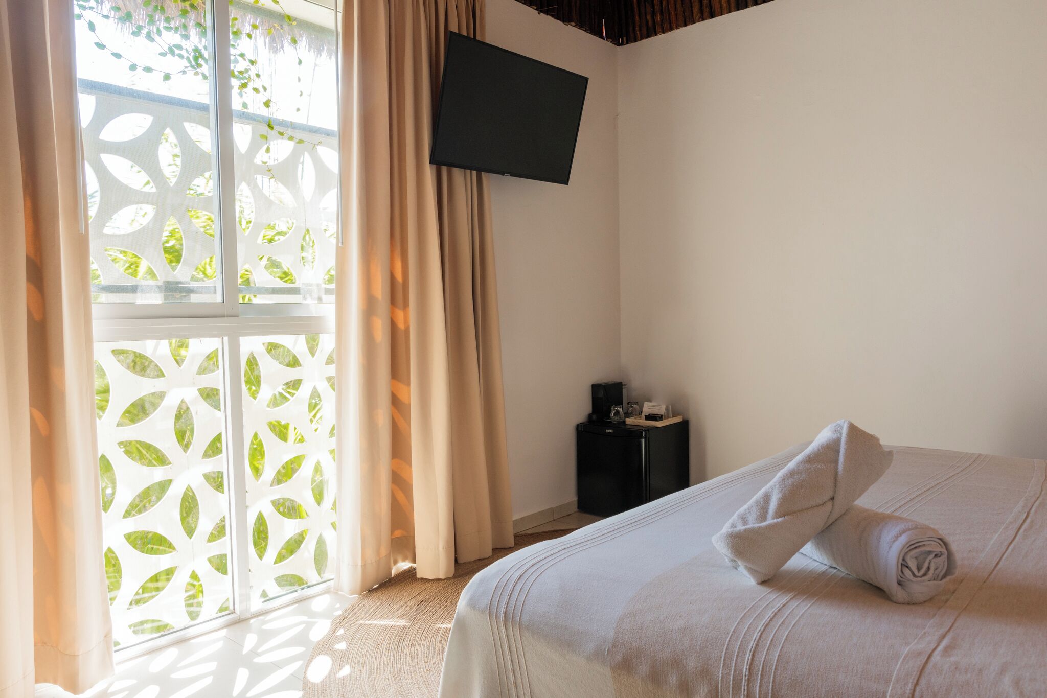 Une chambre d’hôtel comprenant un lit, un téléviseur à écran plat fixé au mur, une petite table de chevet noire et une fenêtre agrémentée d’un paravent décoratif.