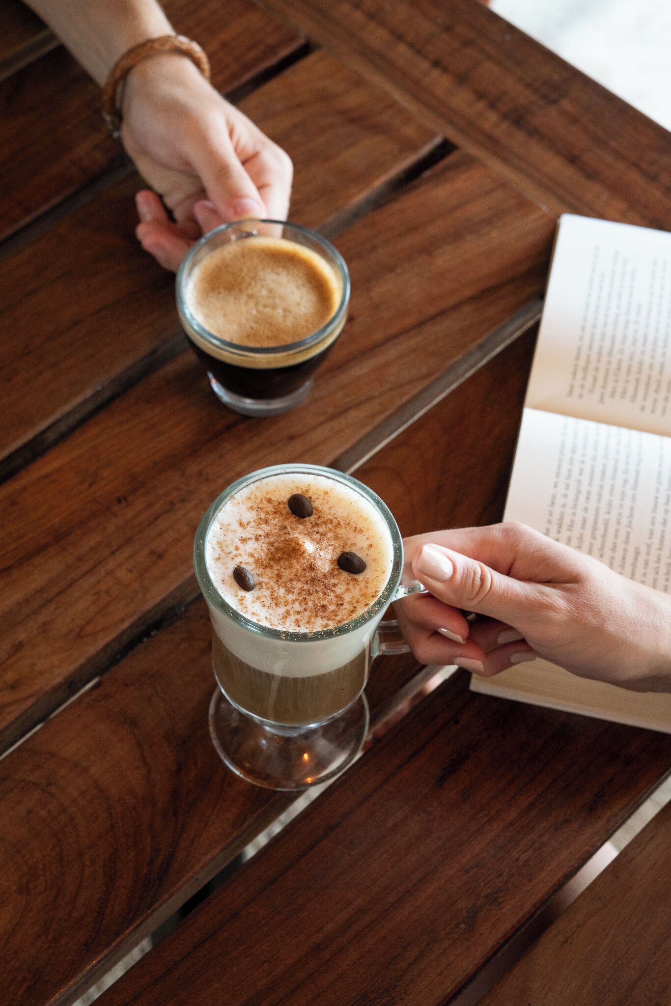 Two hands holding a cup of coffee with a frothy top and a sprinkle of cinnamon, and a book on a wooden table.