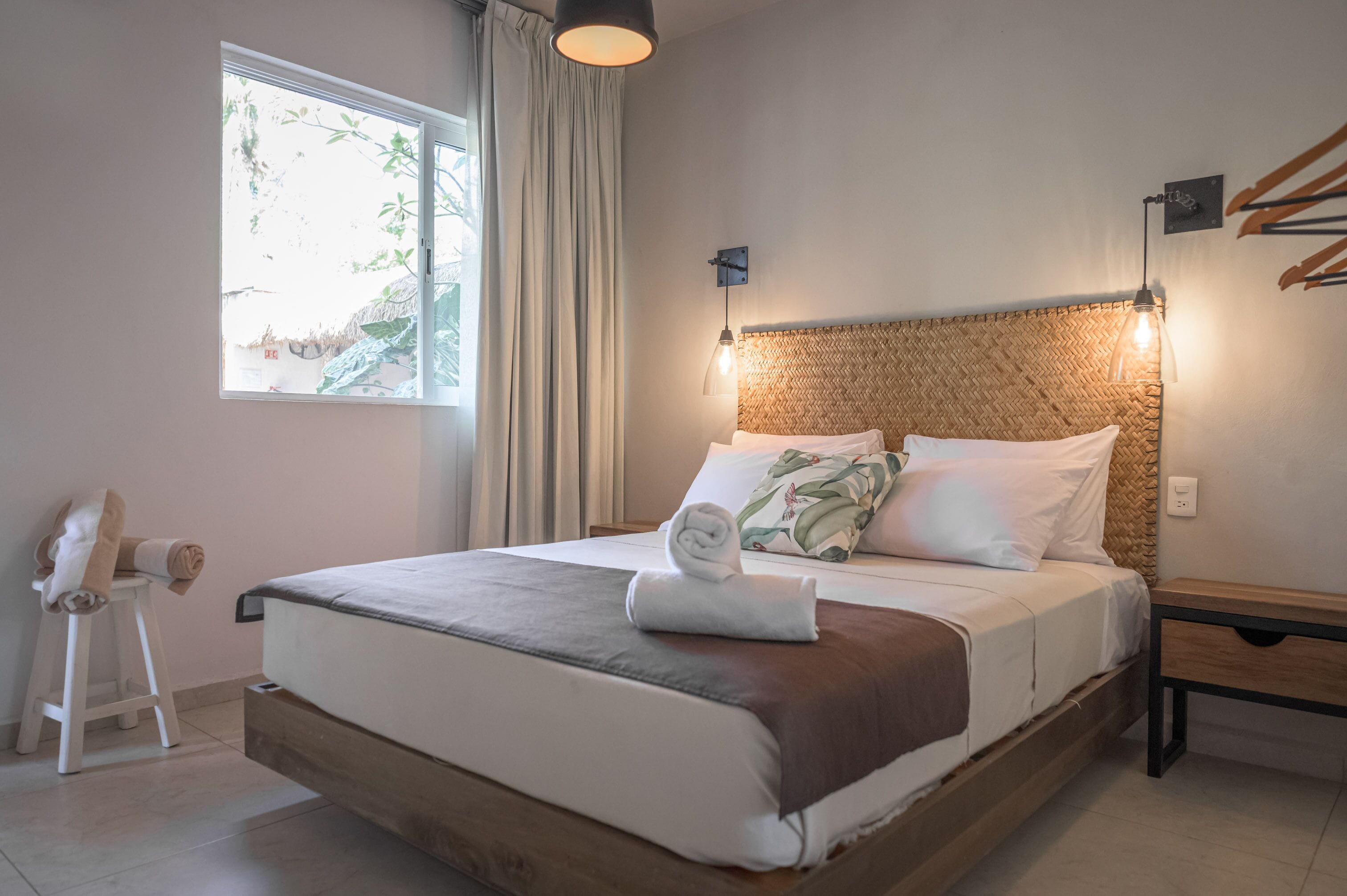 Una cama bien hecha con cabecera marrón y un amplio ventanal con cortinas.