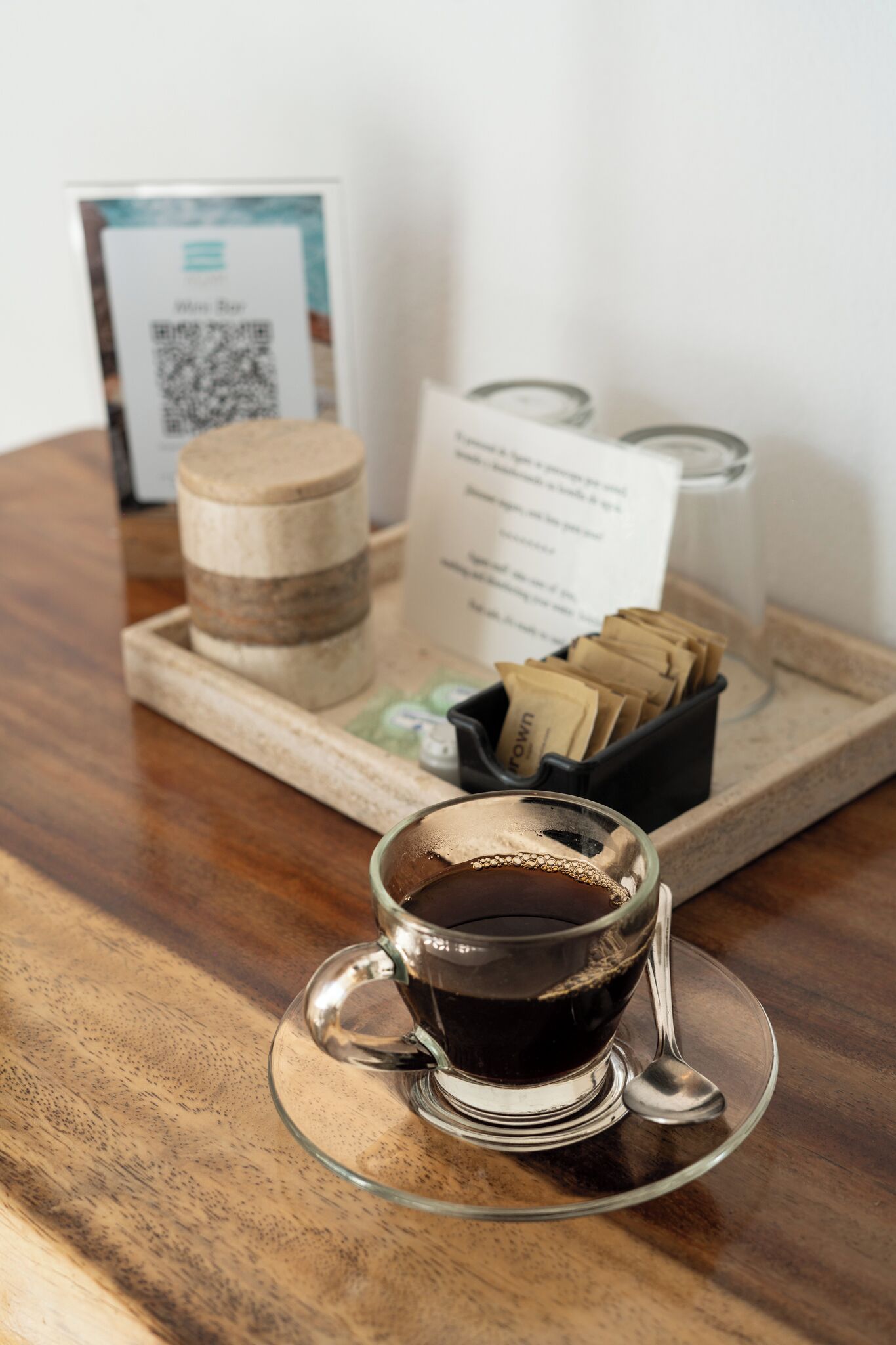Une tasse de café en verre, posée sur un plateau avec une cuillère, un pot et un panneau avec un code QR.