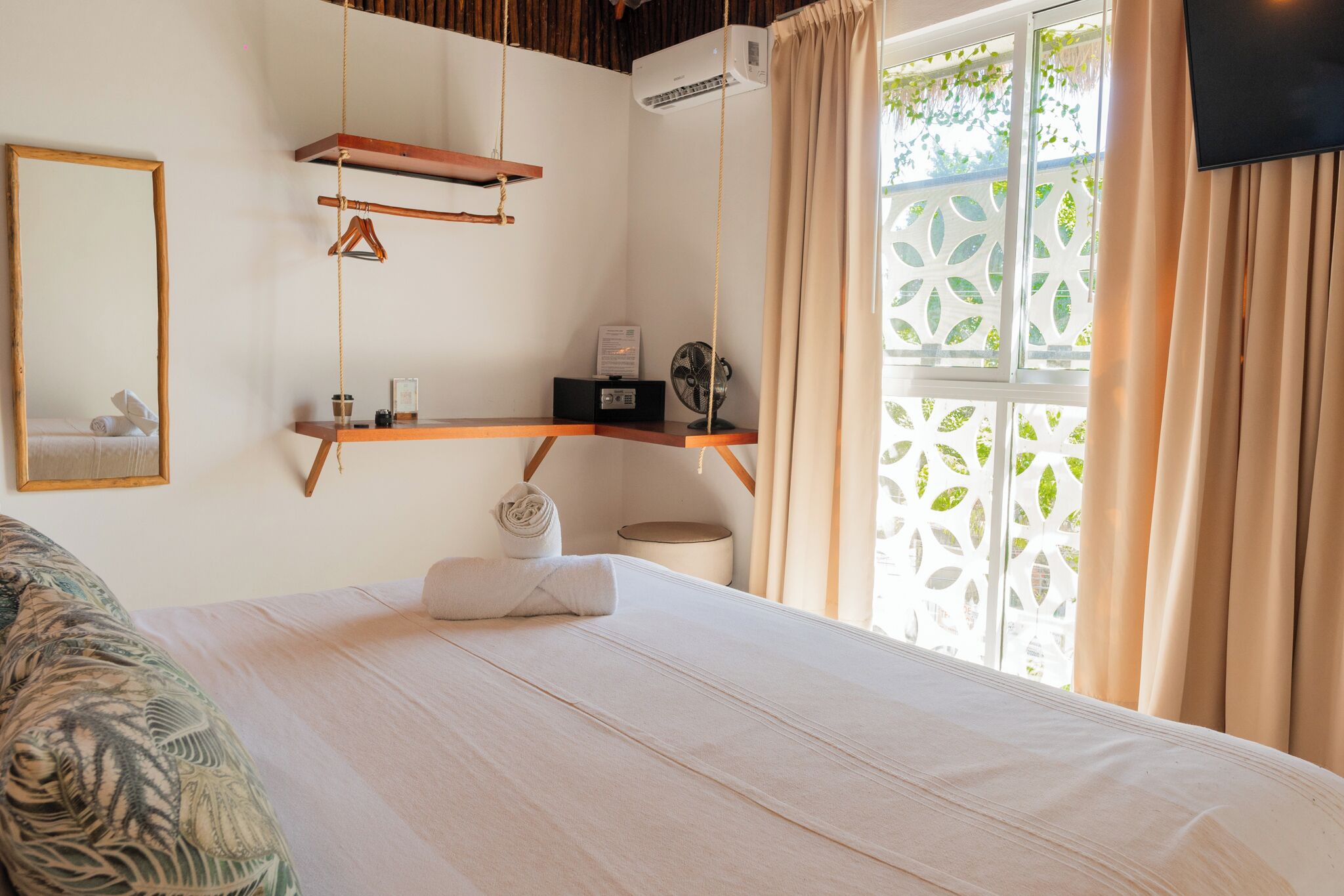 A bedroom with a bed, a hanging chair, a small table with a fan, and a window with curtains.