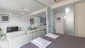 Ein modernes Schlafzimmer mit einem großen Bett, einem Sofa, einem Essbereich und einer durch eine Glastrennwand sichtbaren Küche.