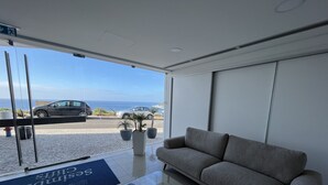 Ein modernes Wohnzimmer mit einer Couch, einem Glastisch und Blick auf das Meer sowie auf parkende Autos durch große Fenster.