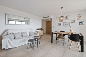 Ein modernes Wohnzimmer mit Sofa, Couchtisch, Esstisch und Stühlen. An der Wand hängt ein großer Spiegel, eine Pendelleuchte und mehrere gerahmte Bilder.