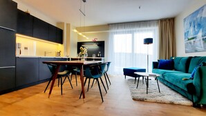 Una sala de estar moderna con un sofá verde azulado, una mesa de comedor de madera y una cocina con armarios oscuros y encimera clara.