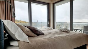Un dormitorio con una cama grande, vistas de la ciudad a través de ventanas de piso a techo y un balcón con mesa y silla.