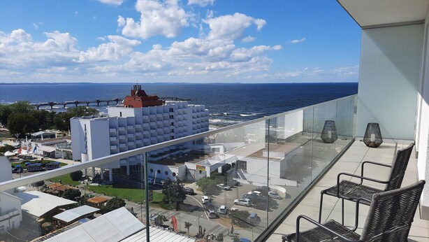 Un balcón con vista al mar, un edificio blanco con techo rojo y un estacionamiento con coches.
