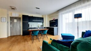 Una sala de estar moderna con piso de madera, un comedor con mesa y sillas, una cocina con gabinetes oscuros y un ventanal con cortinas de gasa.