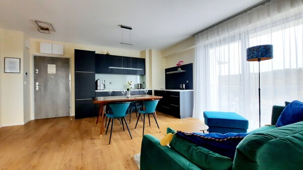Un salon moderne avec un plancher en bois, un coin repas avec une table et des chaises, une cuisine avec des meubles sombres et une grande fenêtre ornée de rideaux légers.